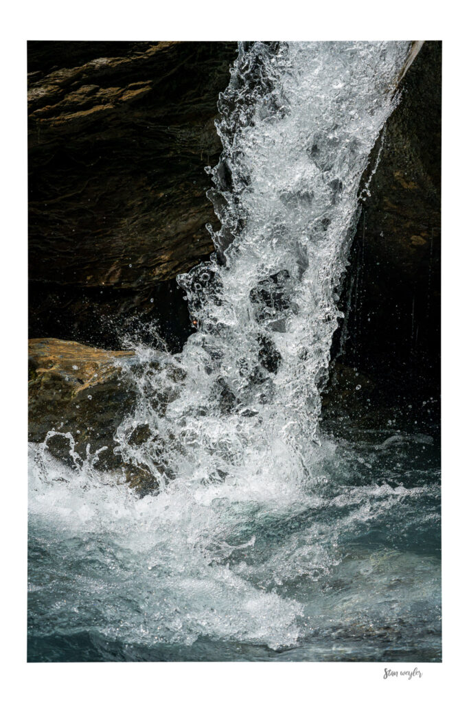 Frozen Waterfall Wall Art, Earth Tone Nature Photography Print by Stan Weyler Art Fine art color photograph of a falling mountain water jet captured with a short time exposure by Stan Weyler. High-speed detail study showing a frozen column of splashing water droplets in front of a dark, mossy rock wall. Minimalist earth-tone nature wall decor for modern interiors, living rooms, and offices, printed on museum-quality archival paper.