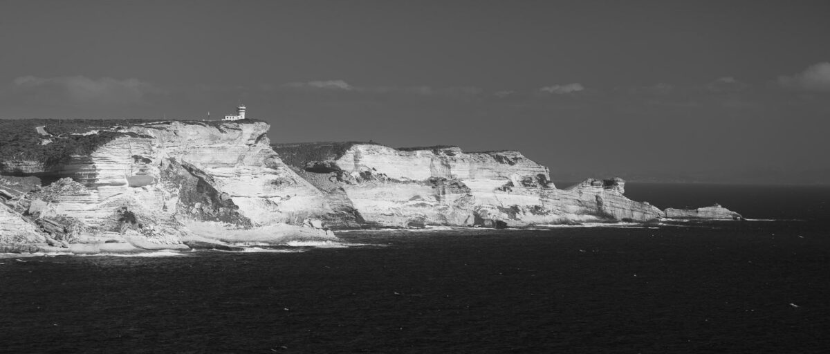 Bring the rugged, dramatic beauty of the Mediterranean into your home with this striking black-and-white fine art print. This photograph captures the iconic Falaises de Bonifacio—the spectacular, chalk-white limestone cliffs on the southern coast of Corsica, France. Towering up to 70 meters above the sea, these sheer drops have been sculpted by centuries of wind and waves along the Strait of Bonifacio. This specific composition takes a minimalist approach, focusing on the southern tip of the massive cliffs as they meet the vast, wind-blown sea under a wide, clear sky. Rendered in deep black and white, the image strips away distractions to emphasize the raw texture, scale, and quiet majesty of this world-famous landmark.
