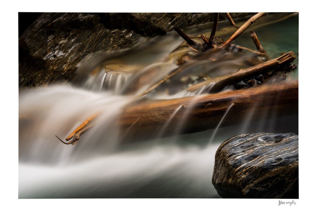 Alpine Driftwood Fine Art Print, Swiss Alps Mountain Stream Photo, Long Exposure River Wall Art, Serene Wilderness Nature Photography by Stan Weyler Art Long-exposure fine art photography print of weathered driftwood wedged between dark rocks in a Swiss Alps mountain stream. High-altitude glacial meltwater flows around the detailed wood, creating a silky, ethereal blur. The wet stones glisten from the spray, showing rich textures. The print features an integrated white gallery border with the photographer's signature 'Stan Weyler' in the bottom right corner.