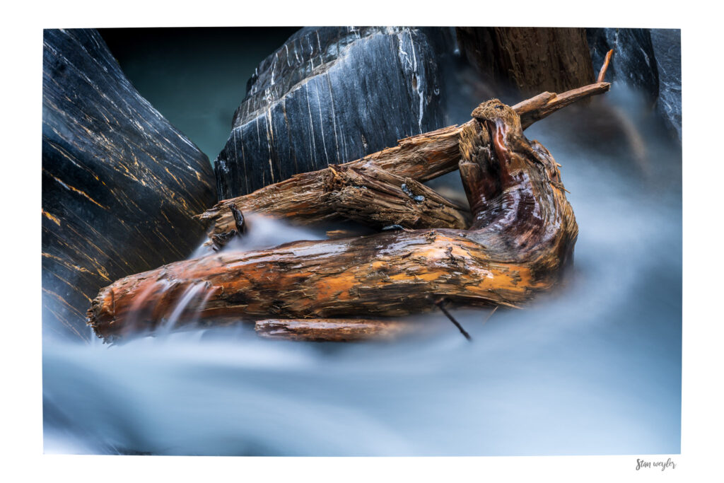 20220505_TIM3473-2-2_Alpine Driftwood Fine Art Print, Swiss Alps Mountain Stream Photo, Long Exposure River Wall Art, Serene Wil Long-exposure fine art photography print of weathered driftwood wedged between dark rocks in a Swiss Alps mountain stream. High-altitude glacial meltwater flows around the detailed wood, creating a silky, ethereal blur. The wet stones glisten from the spray, showing rich textures. The print features an integrated white gallery border with the photographer's signature 'Stan Weyler' in the bottom right corner.
