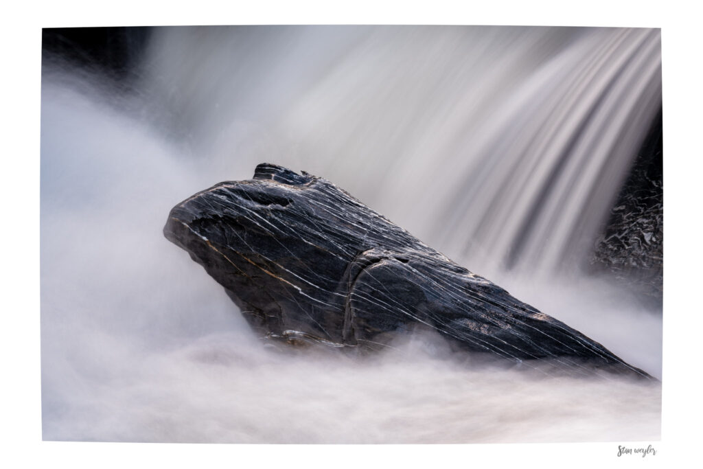 Swiss Alps Long Exposure Waterfall Print, Ethereal Mountain Stream Wall Art Long-exposure fine art photography of a striking dark rock with white quartz veins rising from misty water in a Swiss Alps river. The rock resembles the majestic profile of an eagle's head emerging from a sea of clouds. In the background, a silky waterfall flows seamlessly, creating an ethereal, dreamlike atmosphere. The print features an integrated white gallery border with the artist's signature 'Stan Weyler' in the bottom right corner.