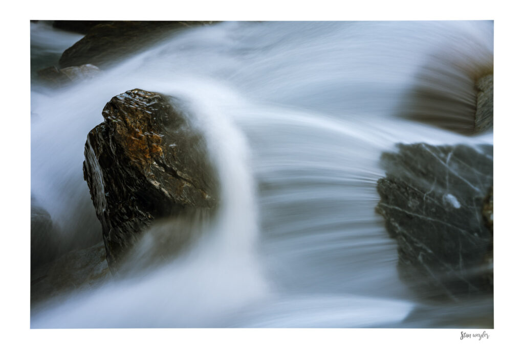 Swiss Alps River Fine Art Print, Long Exposure Water Wall Art, Dark Rock Nature Photography, Ethereal Mountain Stream Gallery Artwork Long-exposure fine art photography of a powerful alpine stream in the Swiss Alps. Silky, linear patterns of rushing water flow dramatically around a dark, wet boulder. The top of the rock glistens with warm, golden undertones from the river spray, contrasting against the cool blue-grey tones of the torrent. The print features an integrated white gallery border with the artist's signature 'Stan Weyler' in the bottom right corner.