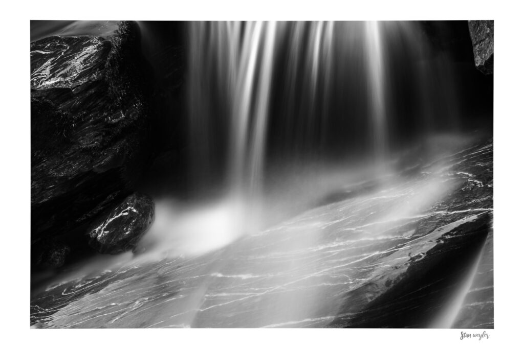 Black and White Waterfall Fine Art Print, Long Exposure River Photography, Swiss Alps Nature Artwork by Stan Weyler Art Black and white long-exposure fine art photography print of an alpine waterfall in the Swiss Alps. Silky vertical light streaks of water cascade down onto a dark, wet rock with prominent white quartz veins. The contrast between deep shadows and smooth white water creates a serene, cosmic, and ethereal atmosphere. The print features an integrated white gallery border with the artist's signature 'Stan Weyler' in the bottom right corner.