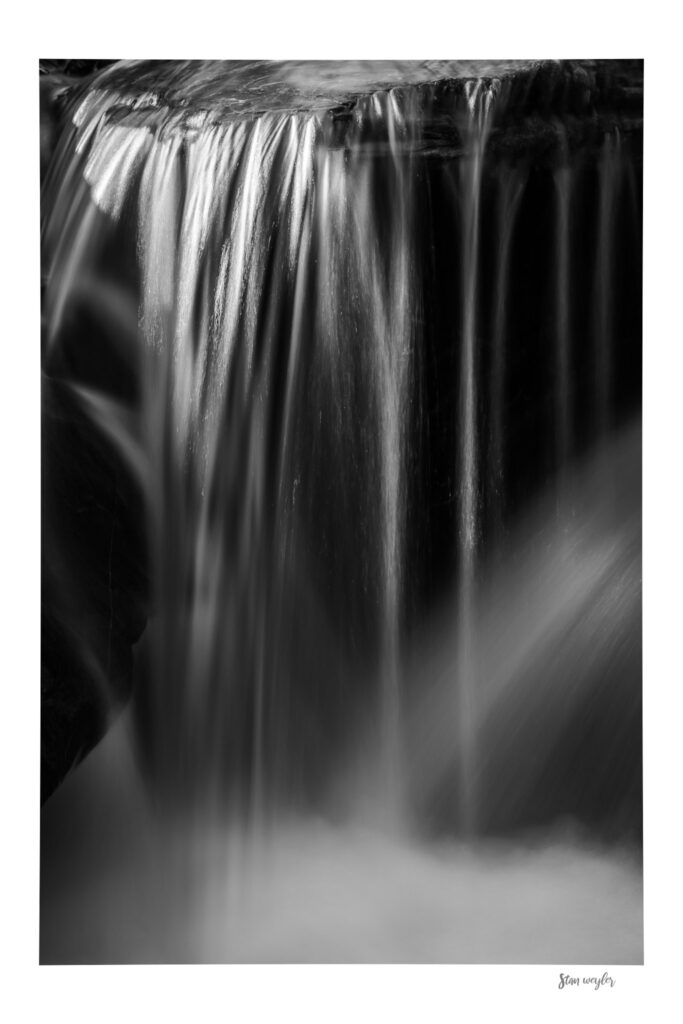 Black and White Waterfall Print, Long Exposure Swiss Alps Nature Photography by Stan Weyler Art Vertical black and white long-exposure fine art photograph of a mountain waterfall in the Swiss Alps. Silky streaks of cascading water shimmer with bright sunlight reflections as they flow over a dark, wet rock edge. A soft diagonal beam of light illuminates the mist below, creating a tranquil and timeless pattern. The print features an integrated white gallery border with the artist's signature 'Stan Weyler' in the bottom right corner.