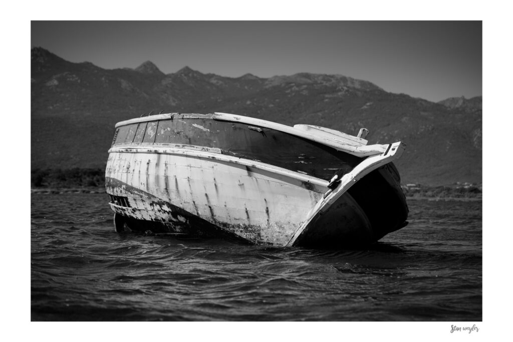 Minimalist Monochrome Coastal Print, Abandoned Boat Photo, Rustic Ocean Textures Wall Art by Stan Weyler Art A rustic and modern maritime fine art photograph of an abandoned, decomposing wooden boat hull in a shallow bay near Bonifacio. Shot from a low angle at the water's surface, highlighting the textures of peeling white paint and vertical erosion marks on the wood. The high-contrast print features a white mat border, an artist signature by Stan Weyler Art.