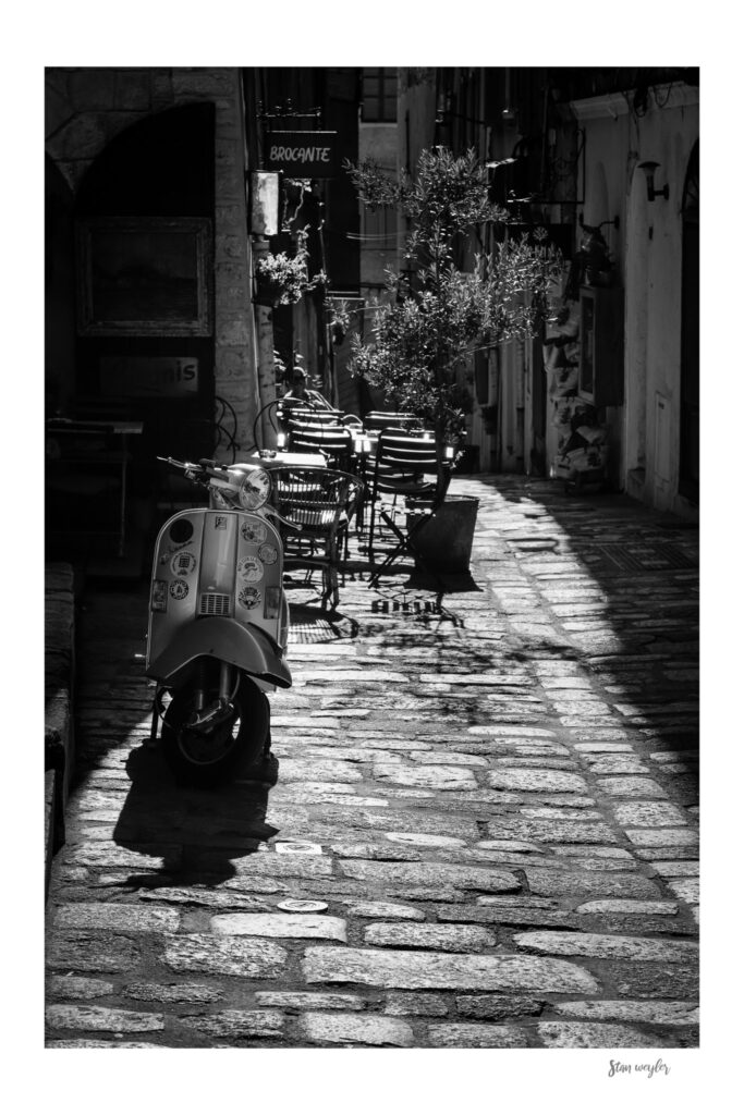Vintage Vespa Print, Bonifacio Corsica Black White Photo, Mediterranean Alley Street Decor (Signed by Stan Weyler) A black and white photograph of a vintage scooter parked on a cobblestone street. The scooter features multiple circular stickers on its front panel. In the background, outdoor cafe seating is visible near a sign reading BROCANTE (Signed by Stan Weyler).