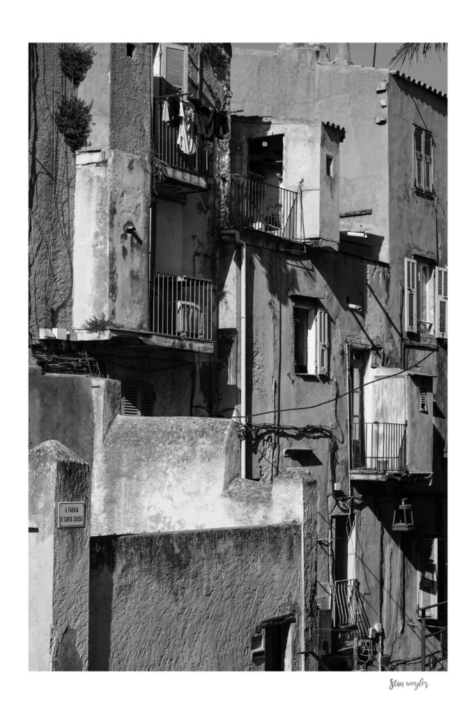 MIDDAY GEOMETRIES - Discover the hidden graphic poetry of a historic Mediterranean old town. This captivating black and white fine art photograph explores the intricate, interlocking geometries and weathered textures of an ancient citadel facade. Shot under dramatic side lighting, the composition thrives on sharp, deep shadows and brilliant highlights that bring every detail to life&mdash;from the peeling plaster and rustic window shutters to the charming balconies and drying laundry. Stan WeylerArt.