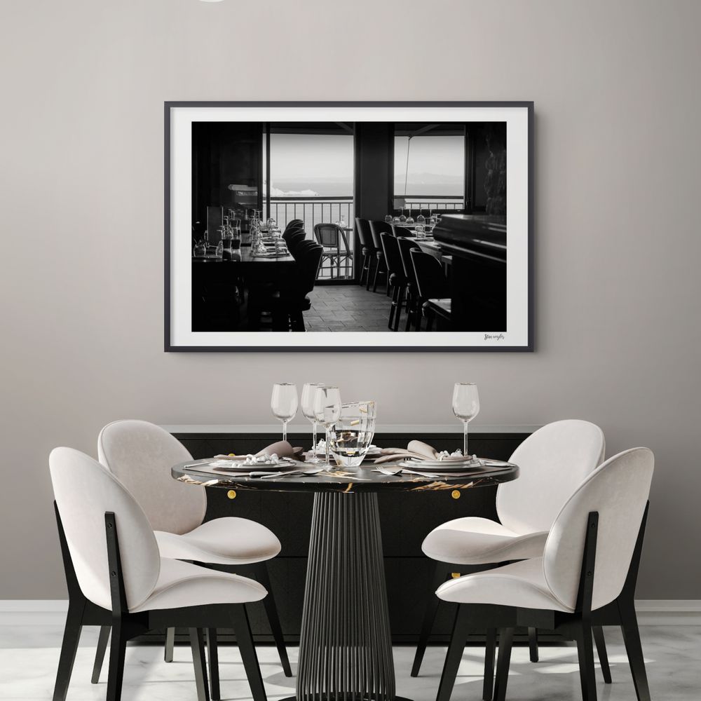 THE STORY BEHIND THE PRINT: „An Hour Before Dinner“ Step into a moment of pure, cinematic suspense high above the Mediterranean. This captivating black and white photograph captures the quiet, moody interior of an empty bistro inside the ancient Citadel of Bonifacio, just one hour before dinner begins. The room is enveloped in deep shadows, with dark walls and black wooden chairs waiting for the evening guests. In the beautiful backlight, rows of meticulously set wine and water glasses gleam like polished silver, drawing your eyes across the room toward the bright, sunlit terrace. The bistro and its lonely balcony literally hang in the air, perched on the edge of the sheer limestone cliffs. Through the open door, the eastern edge of the cliffs is visible in the distance, cutting into the famous Strait of Bonifacio. A powerful study of light, contrast, and European nostalgia that brings an elegant, mysterious focal point to your walls. ✨ Elevate your space with this timeless black and white fine art print from Stan WeylerArt. Perfectly balancing minimalist aesthetics with high-contrast depth, this piece brings sophisticated gallery vibes into your home.